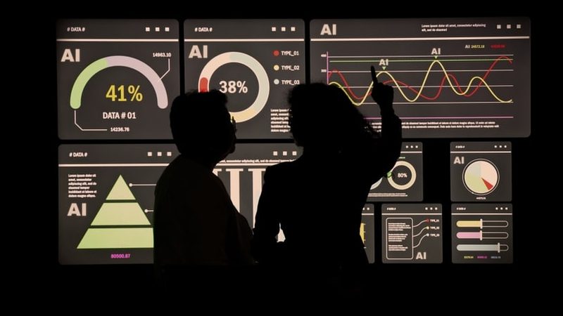 Two people silhouetted against a wall of illuminated AI dashboards and charts