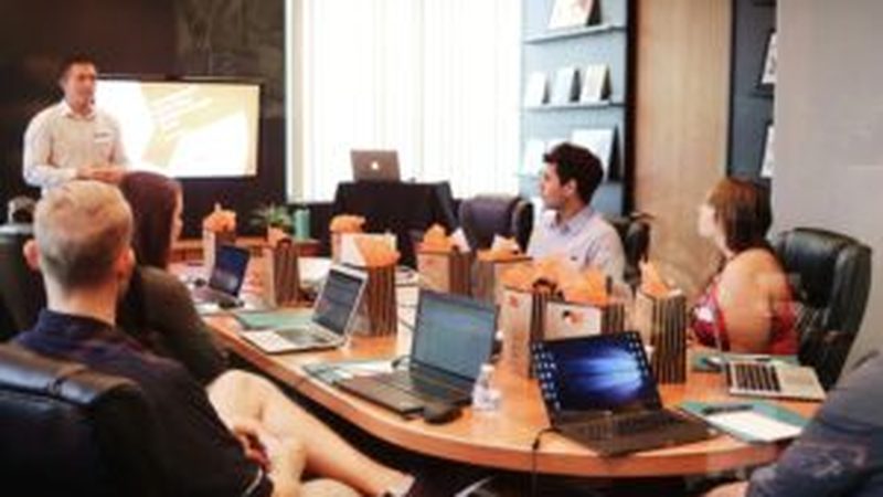 A presenter addressing a group seated around a boardroom table with laptops open