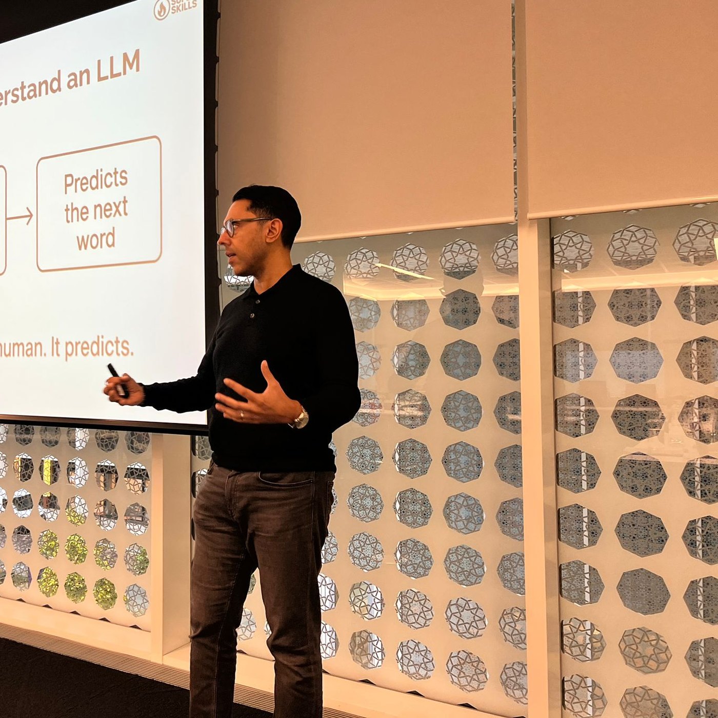 Rahim Hirji presenting to a seated room beside a screen explaining how a large language model predicts the next word