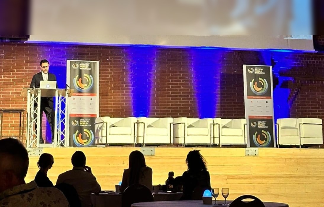 Rahim Hirji presenting from a lectern on the main stage at the EdTech World Forum in London, with a large projection screen above him and the hall lit in blue