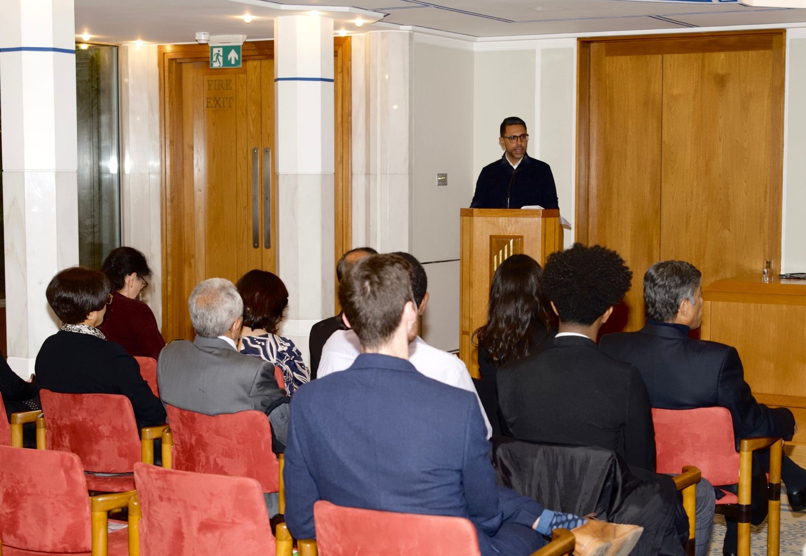 Rahim Hirji speaking from a lectern to a seated audience in a wood-panelled hall