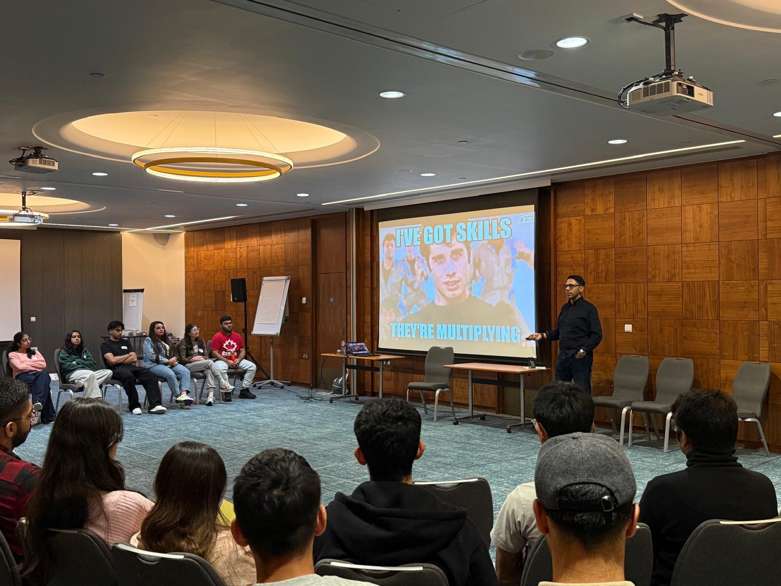 Rahim Hirji standing beside a screen in a room where the audience is seated in a wide circle rather than in rows