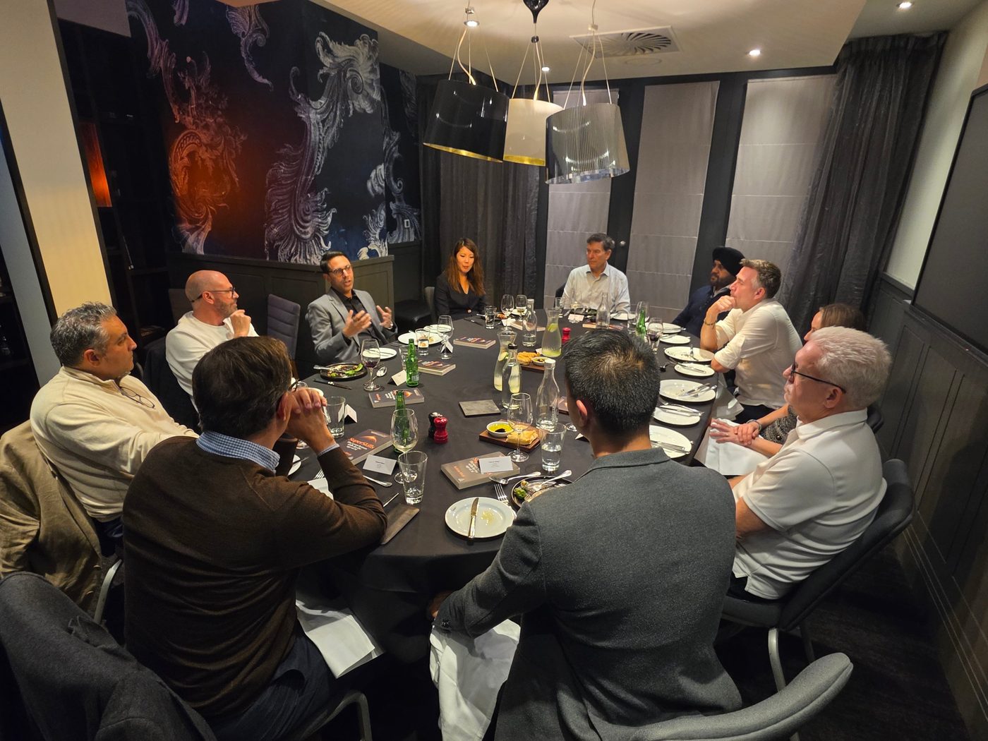 A wide view of a private dining room, Rahim Hirji speaking from one side of a round table of a dozen leaders