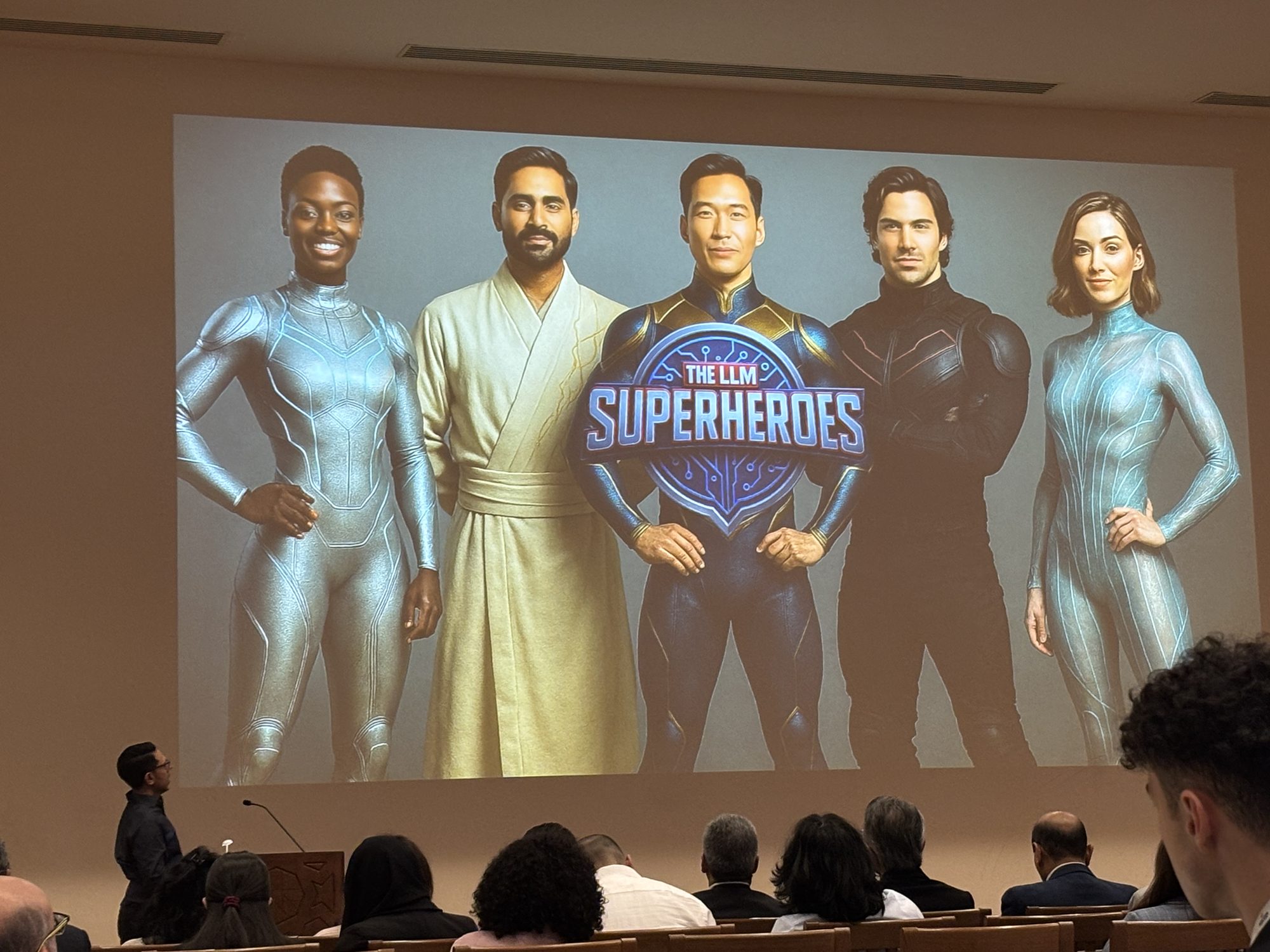 Rahim Hirji presenting from a lectern to a seated audience, with an illustrated slide titled The LLM Superheroes projected behind him